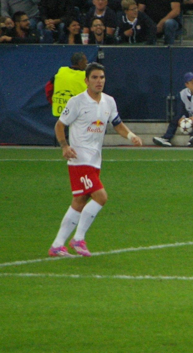 Ein Mann in weißem Hemd und roten Shorts spielt Fußball auf einem Feld, umgeben von einer Gruppe von Menschen im Hintergrund.