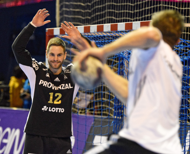 Zwei Männer spielen Handball vor einem Tor mit Netz und einem Banner mit Text im Hintergrund und einigen Menschen in einer Arena.