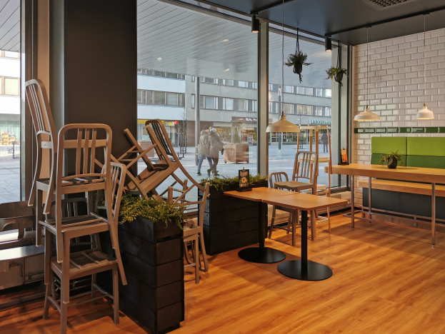 Ein Restaurantinnenraum mit Holztischen und -stühlen in der Nähe eines großen Fensters, das auf eine Berliner Straße mit Fußgängern, Gebäuden und städtischen Elementen blickt.