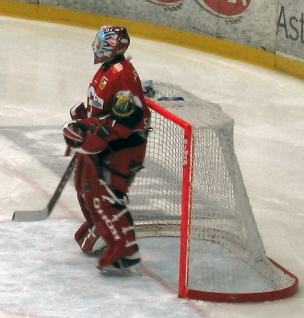 Ein Hockey-Spieler in roter Uniform steht auf dem Eis und hält einen Hockey-Schläger in der Hand und trägt einen Helm, vor ihm steht ein Tor und in Hintergrund eine Wand mit Text.