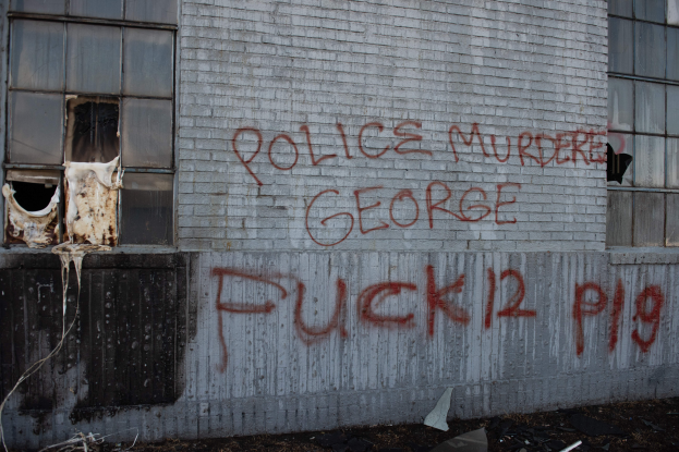 Ein Gebäude mit der Graffiti-Aufschrift "Polizei ermordet George Fuck 12 P19" an der Seite, das Fenster und verschiedene Gegenstände vorne zeigt.