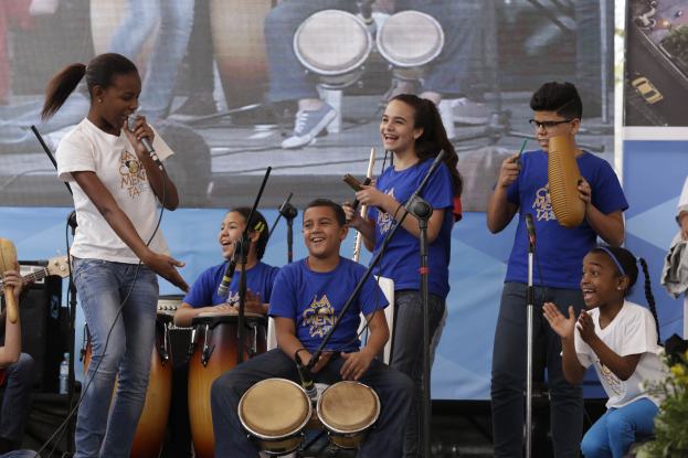 Kinder auf der Bühne mit einer Frau mit Mikrofon, umgeben von Musikinstrumenten und Mikrofonständern, mit einem Banner und Topfpflanzen im Hintergrund.