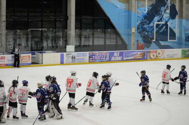Group of young people in helmets and hockey gear playing ice hockey on an indoor rink, with banners, pillars, and glass walls in the background.