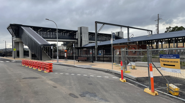 Ein Bahnhofsbild mit einer Brücke im Hintergrund, Straßenmasten, Straßenlaternen, Strommasten, Stromkabel, Bäume, Gebäude, Kraftfahrzeuge auf der Straße, Absperrmasten, Schilder, Bänke und einen bewölkten Himmel.
