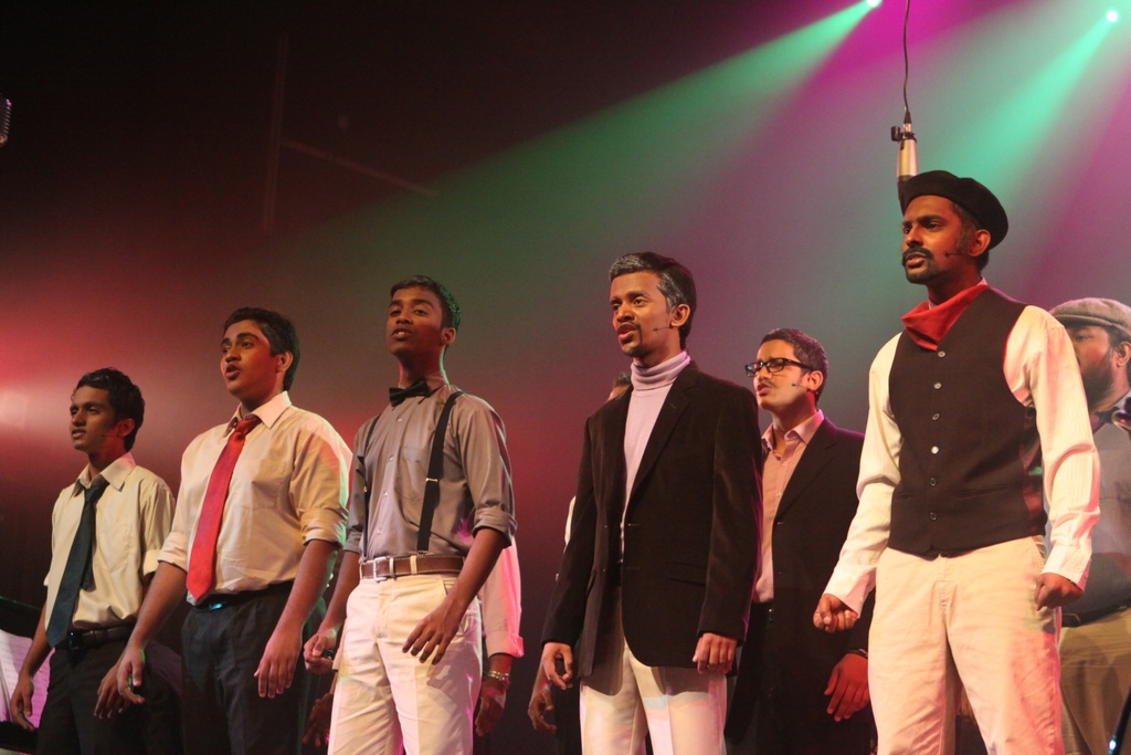 Eine Gruppe von Männern führt ein Drama auf der Bühne auf, im Hintergrund sind grüne Scheinwerfer zu sehen.