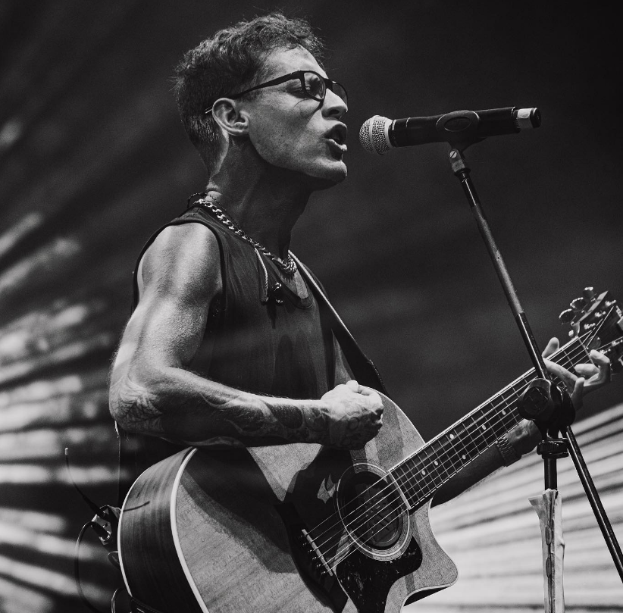 Ein Mann mit Brille spielt eine Gitarre auf der Bühne im O2 Academy Brixton mit einem Mikrofon davor und einer Wand im Hintergrund, dargestellt in Schwarz-Weiß.