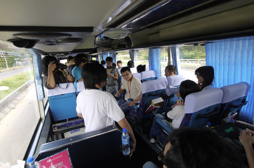 Ein Bus voller Passagiere auf einer Straße, mit einem Zaun auf der linken Seite, einer Straßenabsperrung auf der rechten Seite und Bäumen und Pflanzen im Hintergrund. Einige Gegenstände sind im Bus zu sehen, und eine Person hält eine Kamera. Lautsprecher sind am Dach eines Fahrzeugs befestigt.