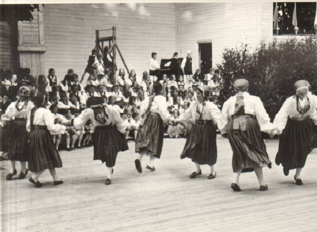Eine Gruppe von Frauen tanzt vor einem gemischten Publikum aus sitzenden und stehenden Menschen, mit einem Gebäude, Pflanzen und einem hölzernen Objekt im Hintergrund, dargestellt in Schwarz-Weiß.