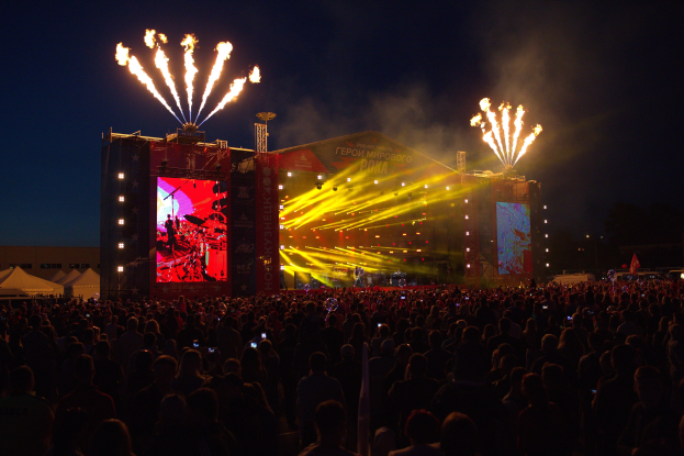 Eine große Menge bei einem Musikfestival schaut auf einer hell erleuchteten Bühne mit Bannern, umgeben von Zelten und Feuerwerk am dunklen Himmel.