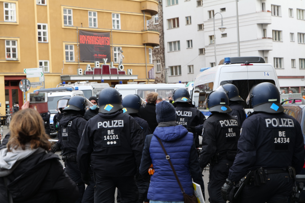 Eine Gruppe von Polizisten steht vor einer Menge von Menschen mit Helmen und Jacken bei einer Demonstration in Berlin, Deutschland, mit Fahrzeugen, Gebäuden, Laternenpfählen und einem Banner mit Text im Hintergrund und einer Person mit einer Kamera.