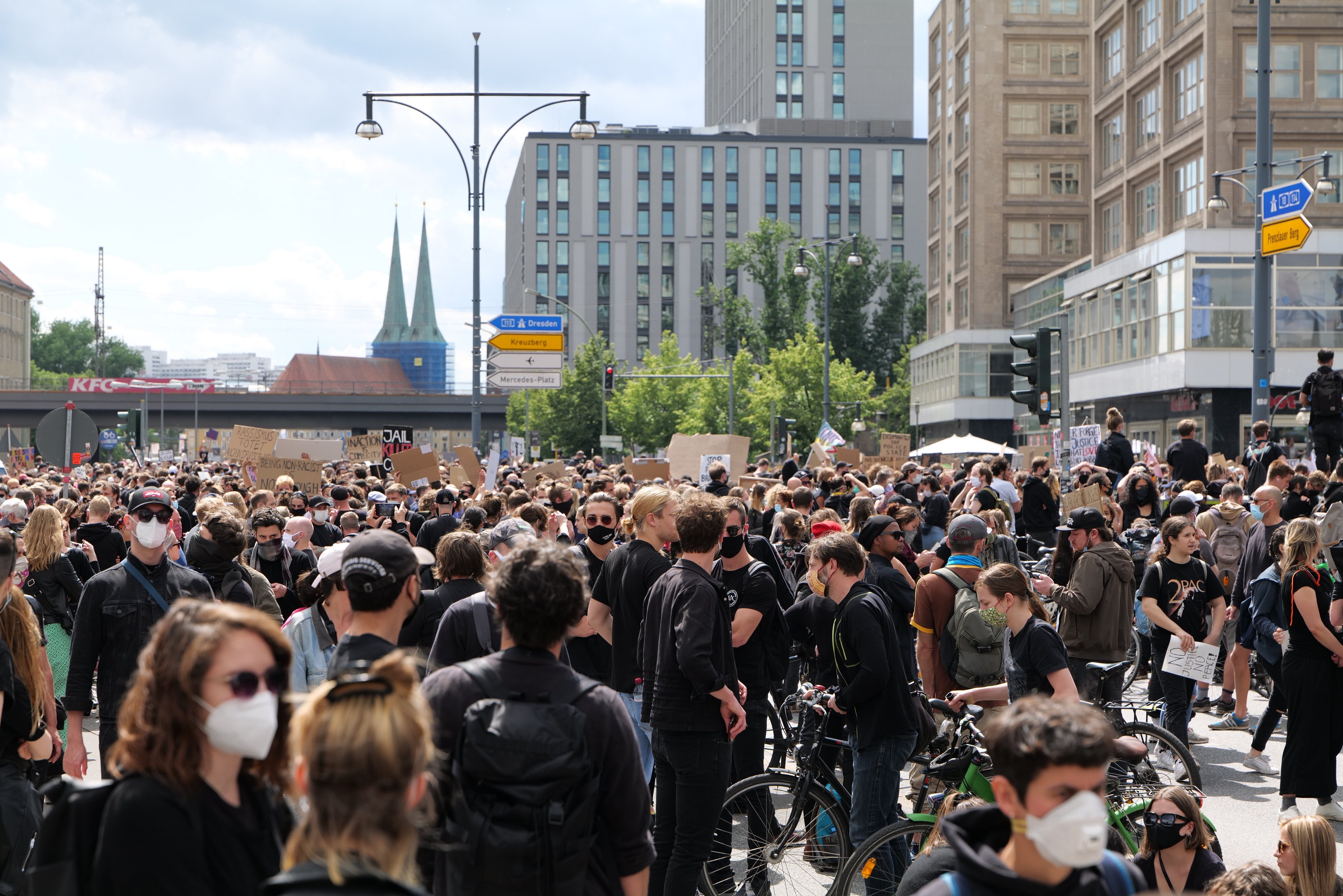 Eine große Gruppe von Menschen mit Masken, die eine von Bäumen gesäumte Straße in Berlin entlanggehen, einige halten Fahrräder, mit Gebäuden, Ampeln und bewölktem Himmel im Hintergrund.