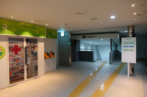 Der Eingang einer Apotheke mit einem Schild, auf dem "Apotheke" steht, auf der linken Seite, einer Tafel mit Text auf der rechten Seite und beleuchteten Lampen auf dem Dach.