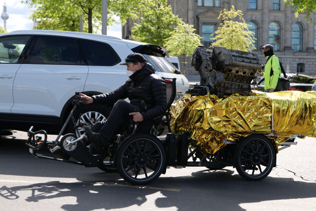 Ein Mann im Rollstuhl mit einem großen Motor auf dem Rücken, umgeben von Fahrzeugen auf einer Straße mit Bäumen, Gebäuden und einem klaren blauen Himmel im Hintergrund; er trägt eine schwarze Jacke, eine Mütze und hält ein Objekt.