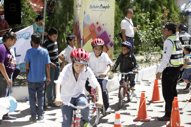 Kinder fahren mit Fahrrädern eine Straße entlang, die von Verkehrskegeln gesäumt ist, einige tragen Helme, andere stehen daneben, mit einem Banner, Bäumen und Gebäuden im Hintergrund.