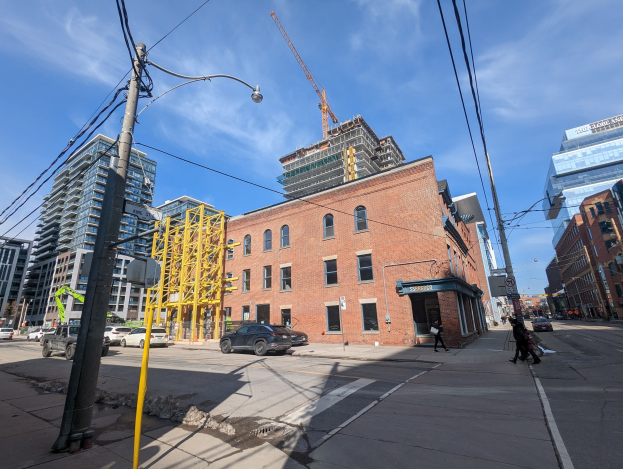 Eine belebte Stadtstraße mit fahrenden Autos, Fußgängern auf dem Gehweg, Strommasten, Straßenlaternen und Gebäuden, darunter ein teilweise errichteter Hochhaus mit einem Kran am Himmel.