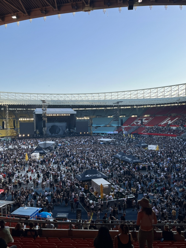 Große Menschenmenge auf Sitzplätzen und Stehplätzen in einem Stadion mit Zelten, Geländern und Überdachung, vor einem Himmel als Hintergrund, was auf ein Konzert in der O2 Arena in London hindeutet.