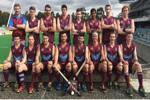 Eine Gruppe von Männern, die auf einem Feld mit Hockey-Schlägern feiern, ein Stadion und Zuschauer im Hintergrund unter einem klaren blauen Himmel.