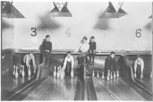 Ein Schwarz-Weiß-Foto von Kindern, die Bowling in einer Gasse mit aufgestellten Kegeln, Zahlen an der Wand und Lampen an der Decke spielen, mit Text unten.
