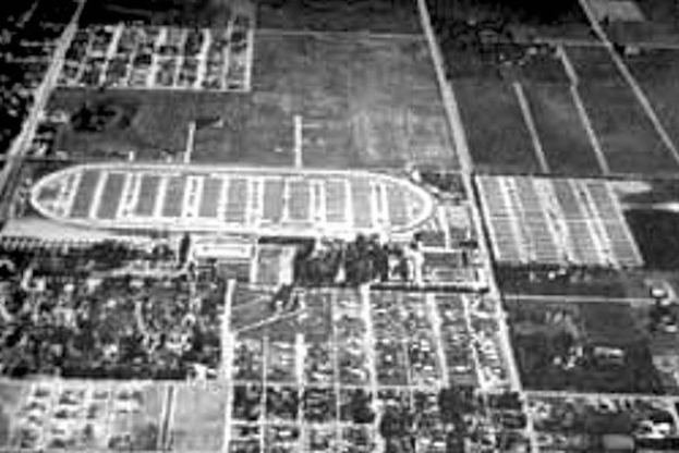 Schwarzer und weißer Luftaufnahme eines Fussballfeldes in städtischer Umgebung, umgeben von Gebäuden, Bäumen und Fahrzeugen.
