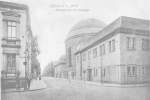 Ein altes Schwarz-Weiß-Foto einer Stadtstraße namens Goethestraße in Offenbach am Main, das Gebäude, Straßenlaternen, Straßenlaternen, Bäume und Menschen zeigt, die auf der Straße gehen, mit dem Text "Offenbach a Main, Goethestraße mit Synagoge" oben.
