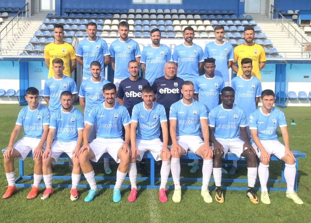 Männer in Sportbekleidung, die auf einem Fußballfeld stehen und sitzen, mit Tribünen und architektonischen Elementen im Hintergrund, wahrscheinlich nach einem Sieg.