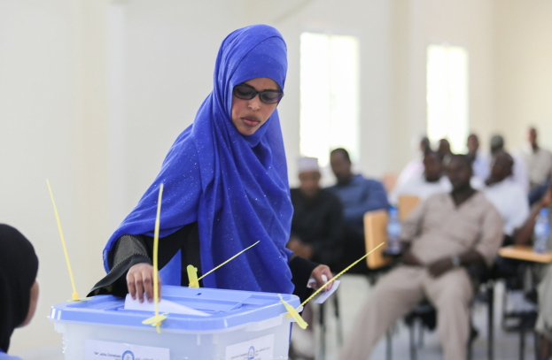 Eine Frau mit einem blauen Kopftuch stimmt in einer Wahllokal ab, während andere daneben sitzen.