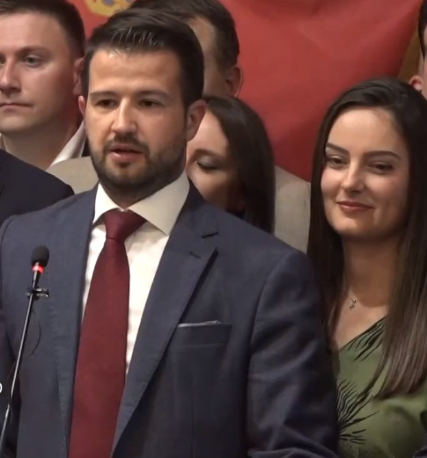 Ein Mann im Anzug spricht an einem Podium mit Mikrofon zu einer Menge hinter einem Banner, wahrscheinlich über ein gouvernementales Verbot von sozialen Medien.