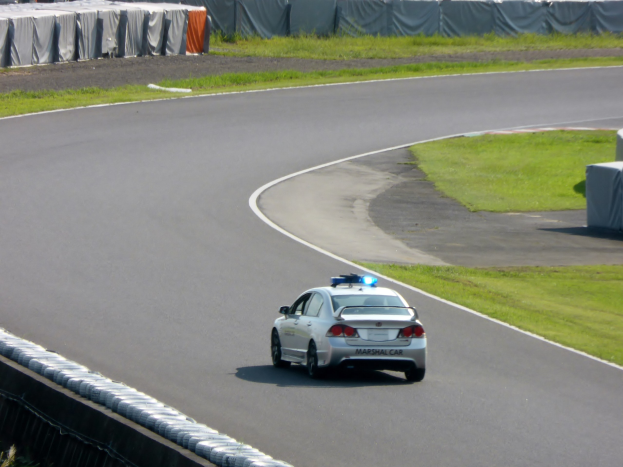 Polizeiauto auf einer Rennstrecke mit Gras auf beiden Seiten und Zuschauern, Zelten und Gegenständen im Hintergrund.