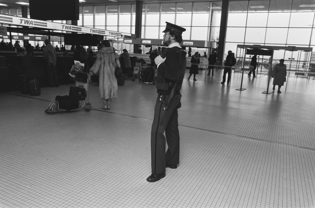Ein Polizist in Uniform steht in einem Flughafen-Terminal und hält eine Waffe in der Hand, umgeben von Menschen und Gepäckwagen, mit Masten, Texttafeln, Deckenleuchten und Glaswänden im Hintergrund.