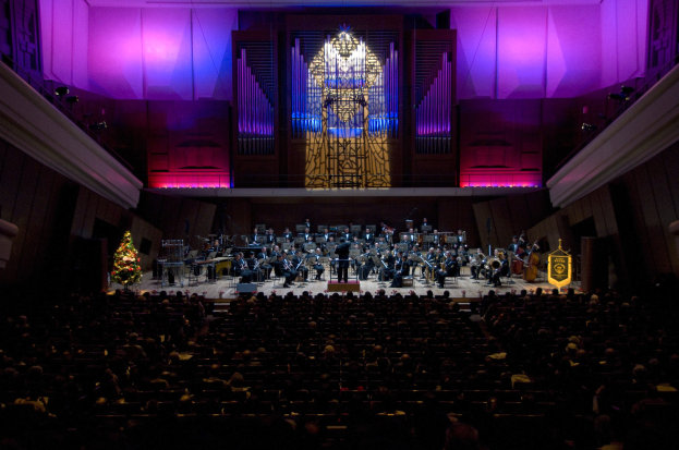 Großer Zuschauerraum voller Menschen bei einem Konzert, mit Musikern auf der Bühne und einem Weihnachtsbaum im Hintergrund.