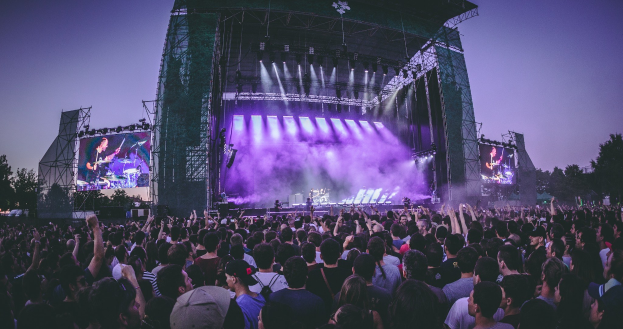 Eine große Menschenmenge auf einem Musikfestival schaut auf einer beleuchteten Bühne mit Instrumenten und Bildschirmen, die vor einem Gebäude, Bäumen und einem klaren blauen Himmel aufgebaut sind.