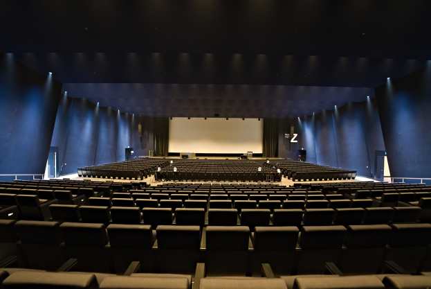 Ein großer Zuschauerraum mit Reihen leerer Stühle, ein zentraler Projektionsschirm und Bühnenobjekte, beleuchtet von Wandlampen.