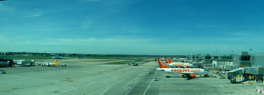 Eine Luftaufnahme eines Flughafens mit Flugzeugen, Gebäuden, Masten, Fahrzeugen, Bäumen, einer Straße unten und einem Zaun im Vordergrund.