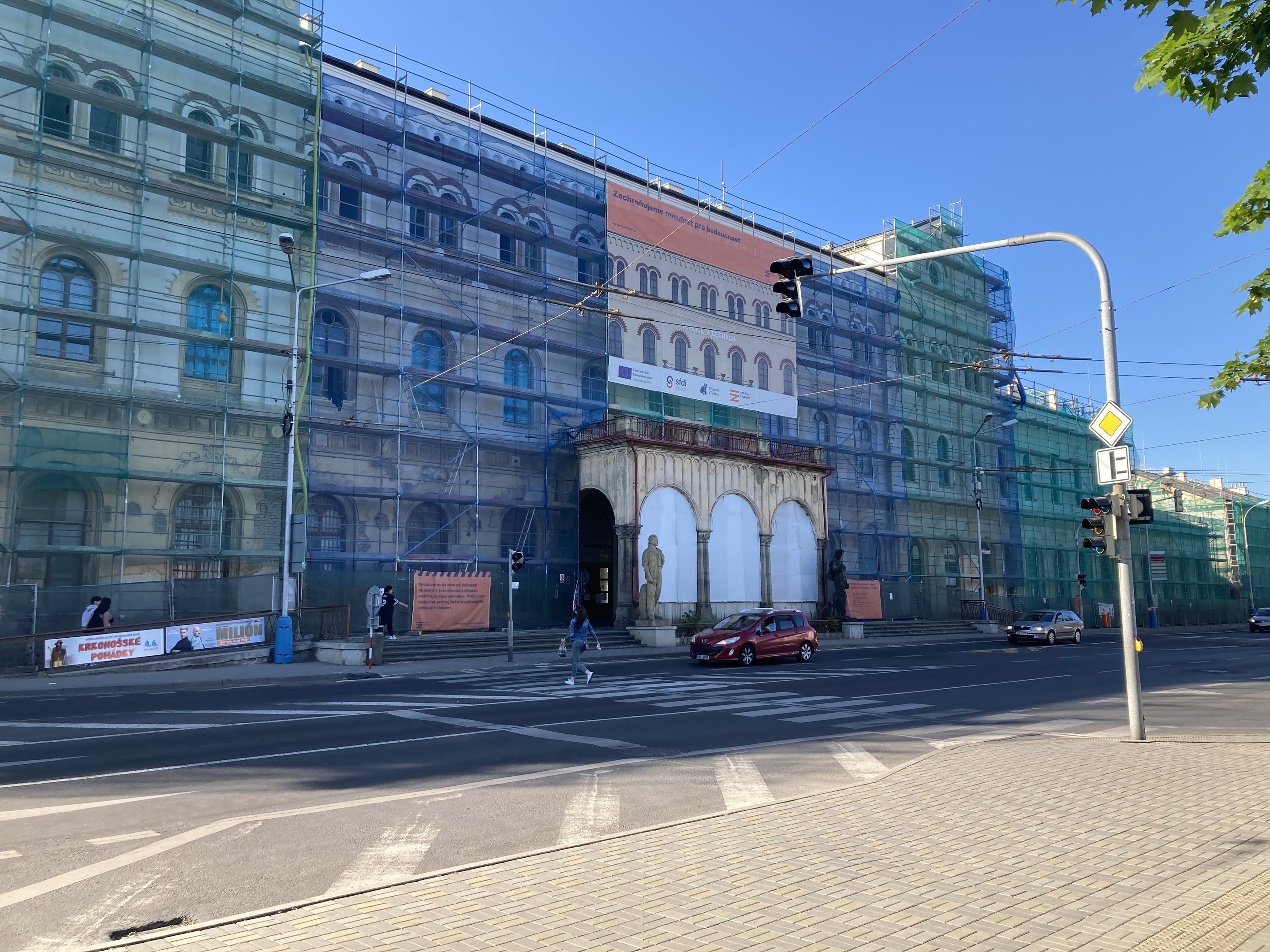 Großes Gebäude mit Gerüst, das renoviert wird, umgeben von Fahrzeugen, Passanten, Ampeln, Schildern, Straßeninfrastruktur, Bäumen und einem klaren blauen Himmel.