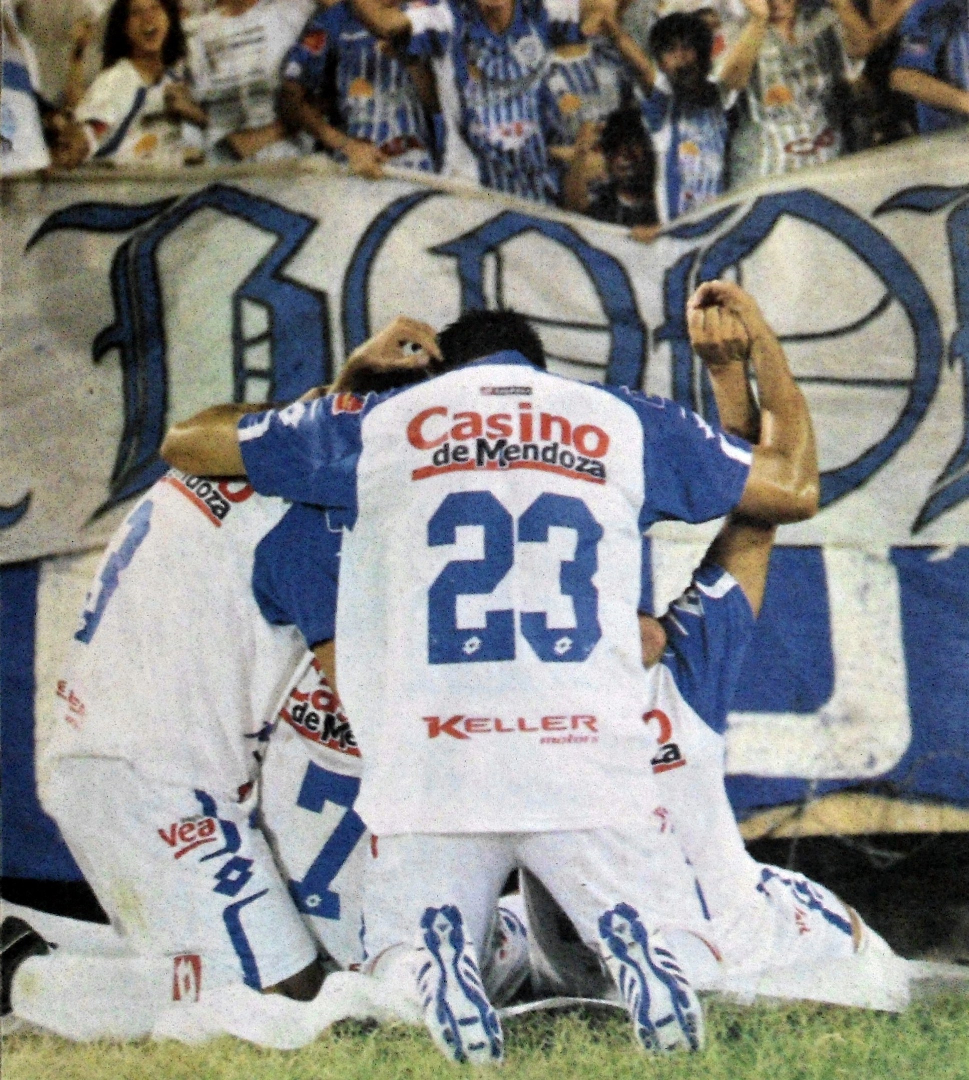 Gruppe von Fußballspielern, die auf dem Boden vor einer jubelnden Menge feiern, mit einem Banner im Hintergrund, auf dem Text steht.