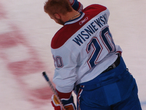 Ein Eishockeyspieler in einem "Wisniewski"-Trikot steht auf dem Eis und hölt einen Eishockeyschläger, mit konzentrierter und entschlossener Miene.