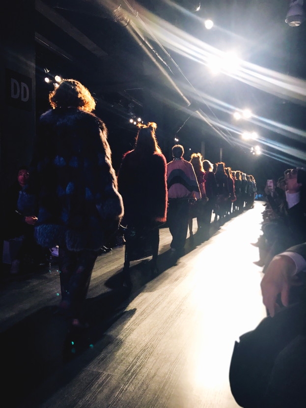 Eine Gruppe von Menschen läuft auf einem Laufsteg bei einer Modenschau, beleuchtet von hellen Scheinwerfern, während Zuschauer auf beiden Seiten die Show beobachten.