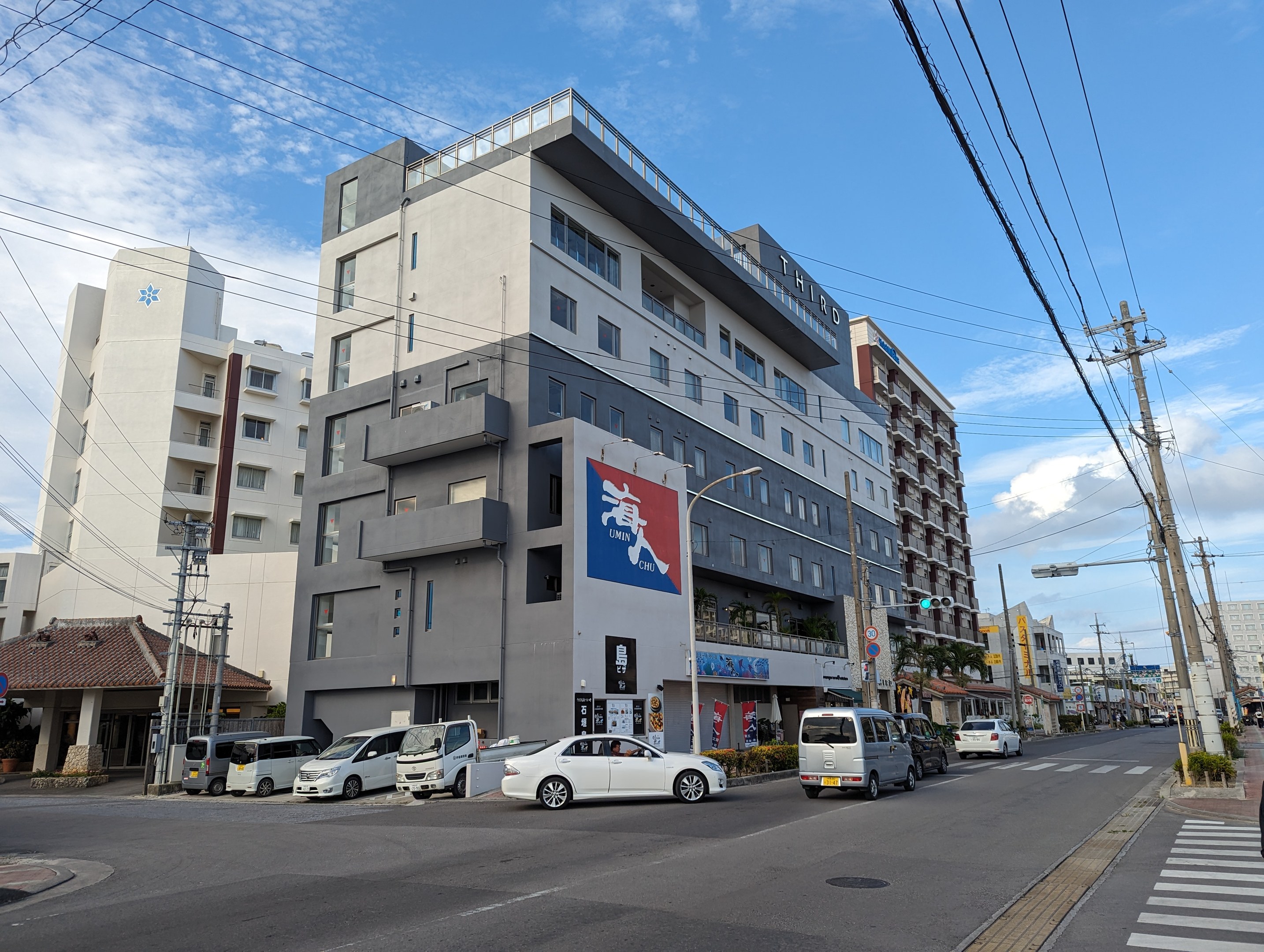 Eine Straße in Nagoya mit parkenden Autos, Strommästen, Laternen und Gebäuden, von denen eines ein "Hotel zum Verkauf"-Schild hat, unter einem bewölktem Himmel.