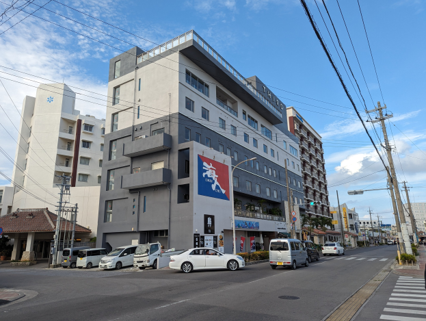 Eine Straße in Nagoya mit parkenden Autos, Strommästen, Laternen und Gebäuden, von denen eines ein "Hotel zum Verkauf"-Schild hat, unter einem bewölktem Himmel.