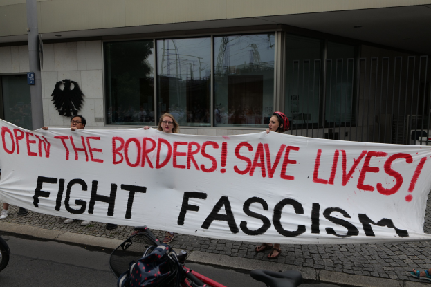 Eine Gruppe von Menschen hält ein Transparent mit der Aufschrift "Öffnet die Grenzen, rettet Leben, kämpft gegen Faschismus" vor einem Gebäude mit Glasfenstern und einer Stange, mit einem Fahrrad und einer Tasche auf der Straße.