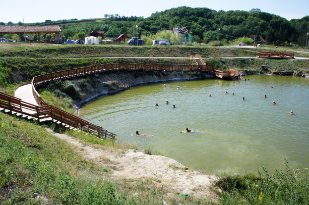 Gruppe von Menschen, die in einem Gewässer umgeben von Grün schwimmen, mit einer Brücke, Treppen, Schuppen, Fahrzeugen, Pfählen und einem klaren blauen Himmel im Hintergrund.
