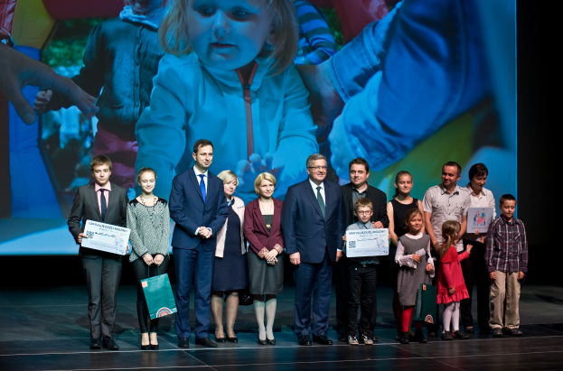 Eine Gruppe von Menschen auf einer Bühne, einige halten Taschen und Urkunden, im Hintergrund ein Bildschirm mit dem Bild eines Kindes und anderer Menschen.