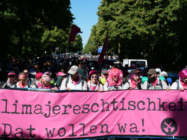 Eine Gruppe von Menschen marschiert mit einem pinken Banner mit der Aufschrift "Frauenmarsch in Berlin" die Straße entlang, einige tragen Möppen und Schutzbrillen, andere halten Fahnen, vor dem Hintergrund von Bäumen, Fahrzeugen und Himmel.