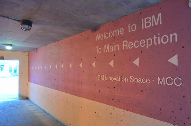 Eine Wand mit der Aufschrift "Willkommen bei IBM", beleuchtet von Deckenlampen, mit einer Tür zum IBM Innovation Space MCC im Hintergrund.