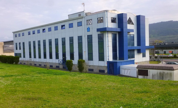 Ein großes weißes und blaues Gebäude mit Fahrzeugen, Masten und Lampen im Vordergrund, vor einer Bergkulisse und einem klaren blauen Himmel.