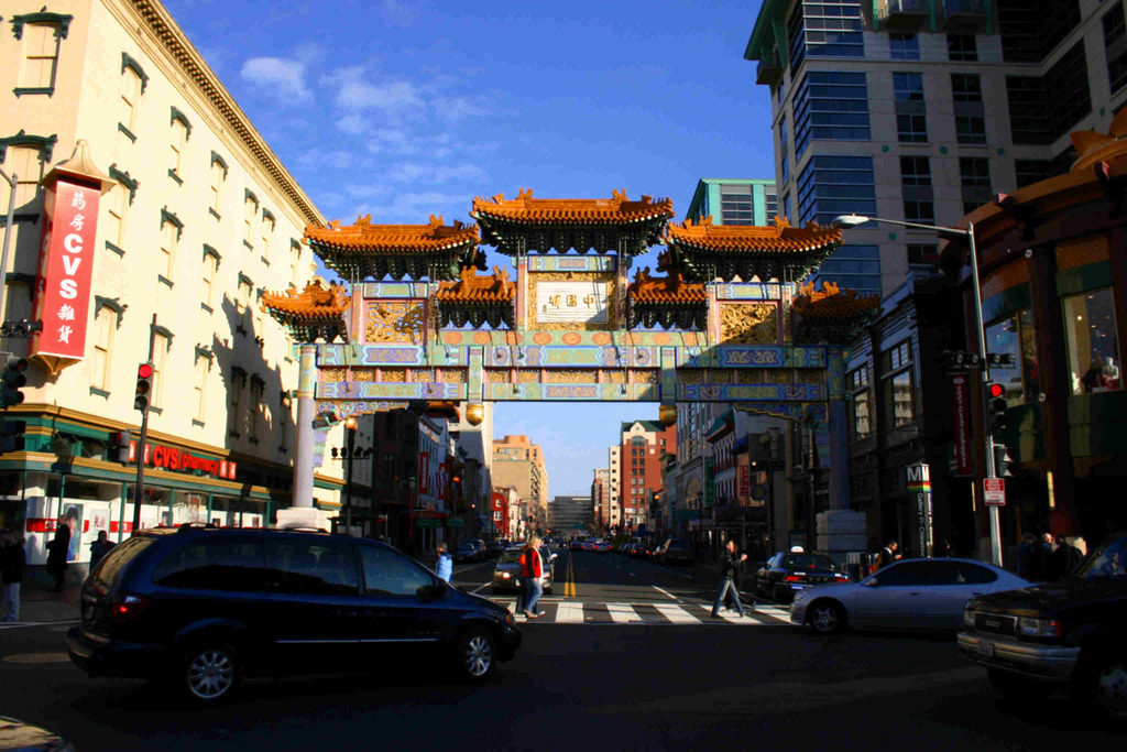 Eine Straße mit fahrenden Fahrzeugen, Fußgänger an einer Zebrastreifen, ein Bogen im Hintergrund, Gebäude auf beiden Seiten und eine Ampel.