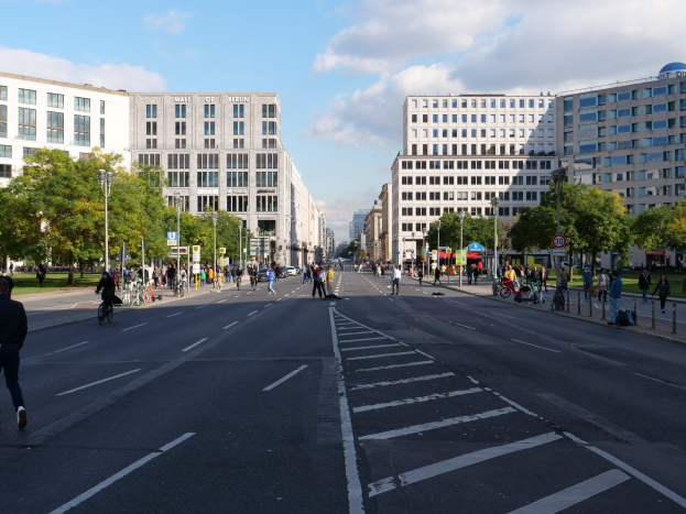 Eine belebte Stadtstraße in Berlin mit Fußgängern und Radfahrern, hohen Gebäuden, Bäumen, Straßenlaternen und Schildern unter einem bewölkten Himmel.