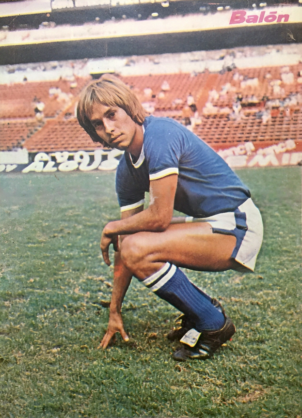 Ein Mann kniet auf einem Fußballfeld, umgeben von Menschen mit Schildern und Sportschuhen.