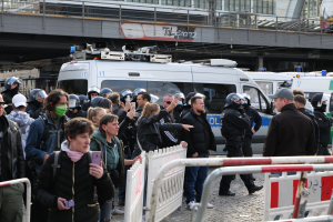 Eine Gruppe von Menschen steht vor einer Reihe von Polizeiwagen in Berlin, Deutschland, einige tragen Helme und halten Mobiltelefone, mit Barrikaden im Vordergrund und einer Brücke mit Geländern und Gebäuden im Hintergrund.