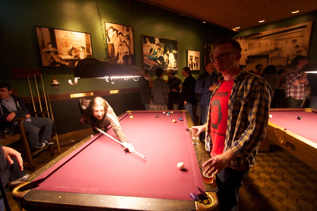 In einem Club spielen zwei Personen Snooker an einem von zwei Tischen, während mehrere andere im Hintergrund stehen und reden, vor einer cremefarbenen Wand, die mit ein paar Fotoschildern geschmückt ist.